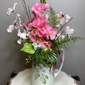 May include: A floral arrangement in a white pitcher with a handle. The arrangement features vibrant pink flowers, white blossoms, and green foliage. The pitcher is decorated with a floral pattern. The arrangement is placed on a white, scalloped-edge plate.