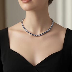 May include: A silver-toned necklace with alternating blue and clear gemstones. The necklace is worn by a person in a black top. The gemstones are evenly spaced, creating a continuous line of sparkle. The necklace is a classic piece of jewellery.