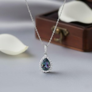 May include: A silver necklace with a pear-shaped pendant. The pendant features a dark teal gemstone surrounded by a halo of small, clear stones. The necklace chain is delicate and hangs on a gray surface.