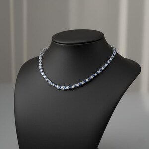 May include: A tennis necklace featuring alternating blue and white gemstones, displayed on a black jewellery bust. The gemstones are set in a silver or white metal setting.