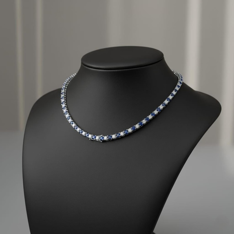 May include: A tennis necklace featuring alternating blue and white gemstones, displayed on a black jewellery bust. The gemstones are set in a silver or white metal setting.