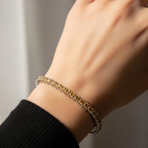 Könnte beinhalten: Ein Tennisarmband aus silberfarbenem Metall mit einer Reihe gelber Edelsteine. Das Armband wird am Handgelenk getragen, wobei ein schwarzes Langarm-Oberteil zu sehen ist. Die Edelsteine sind rund und gleichmäßig angeordnet.