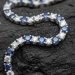 May include: A tennis bracelet featuring alternating blue and clear gemstones set in silver metal. The bracelet is displayed on a black, textured surface. The gemstones are round and evenly spaced.