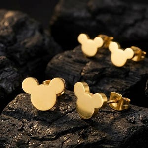 Könnte beinhalten: Goldfarbene Ohrstecker in Form von Mickey Mouse. Die Ohrringe werden auf einer dunklen, strukturierten Oberfläche präsentiert. Sie haben eine glänzende, polierte Oberfläche und ein klassisches Design. Das Bild zeigt mehrere Paar Ohrringe.