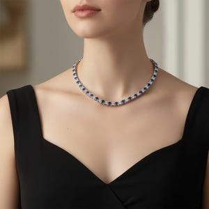 May include: A silver necklace featuring alternating blue and clear gemstones. The necklace is worn around the neck. The gemstones are set in a row. The person is wearing a black dress.