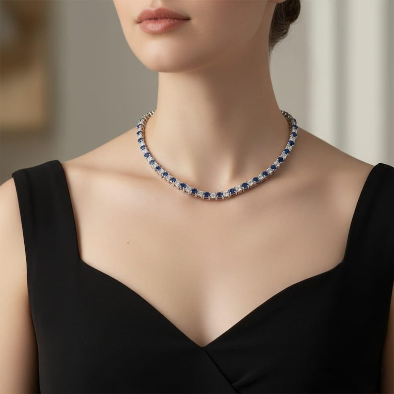 May include: A silver necklace featuring alternating blue and clear gemstones. The necklace is worn around the neck. The gemstones are set in a row. The person is wearing a black dress.