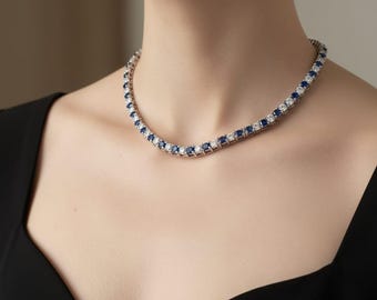 May include: A silver necklace featuring alternating blue and white gemstones. The necklace is worn around the neck, resting on a black top. The gemstones are square-cut and closely set.