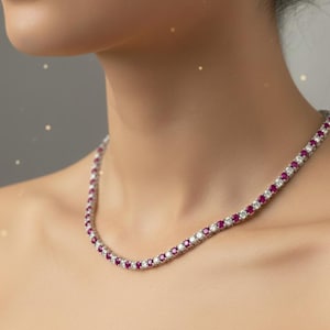 Könnte beinhalten: Eine Tennis-Halskette mit abwechselnd runden Rubinen und Diamanten. Die Halskette ist in einem silberfarbenen Metall gefasst und liegt auf dem Hals einer Person. Die Edelsteine sind leuchtend rot und klar.