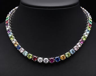 May include: A silver necklace with a line of colourful gemstones. The gemstones are round-cut and set closely together, displaying a spectrum of colours including red, blue, green, yellow, and purple. The necklace is displayed on a black mannequin.