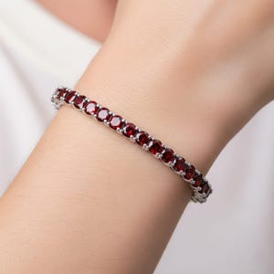 以下が含まれることがあります： 丸い深紅色の宝石が並んだシルバーブレスレット。宝石は互いに密接にセットされ、連続した色のラインを形成しています。ブレスレットは人の手首に装着されています。
