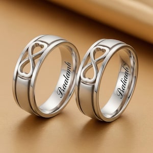 Op de afbeelding: Twee zilveren ringen met een oneindigheidssymbool en hartvorm. De ringen zijn aan de binnenkant gegraveerd met de namen "Raalamli" en "Raliaali". Ze liggen op een goudkleurig oppervlak.