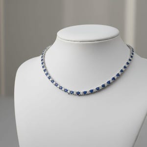 May include: A tennis necklace with alternating blue and clear gemstones. The necklace is displayed on a white mannequin. The gemstones are round-cut and set in a silver-coloured metal. The necklace is a classic piece of jewellery.