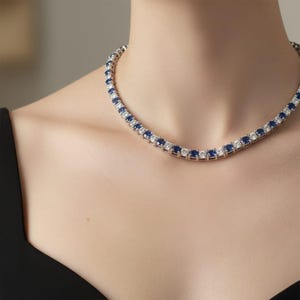May include: A silver necklace featuring alternating blue and white gemstones. The necklace is worn around the neck. The gemstones are square-cut and set closely together, creating a continuous line of sparkle. The necklace is a classic design.