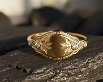 May include: A gold ring with a central oval design featuring leaf engravings and three small diamonds. The ring has a detailed, textured band and is set against a dark, textured surface.