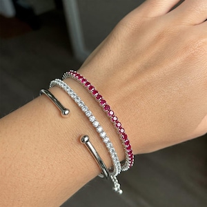 Pode incluir: Três pulseiras em um pulso. Uma pulseira de punho prateada com uma bola em cada extremidade, uma pulseira de tênis com pedras transparentes e uma pulseira de tênis com pedras vermelhas. As pulseiras estão empilhadas.