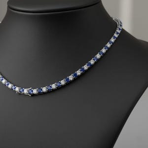 May include: A tennis necklace featuring alternating blue and clear gemstones. The necklace is displayed on a black jewellery bust. The gemstones are round and set in a silver-coloured metal.