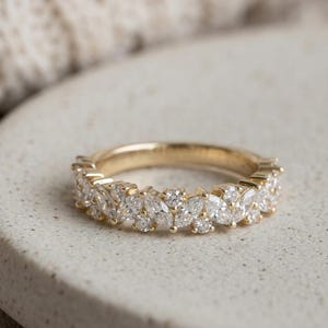 May include: A gold ring featuring a repeating pattern of marquise-cut and round-cut clear gemstones. The ring is displayed on a neutral surface, with a soft, textured backdrop.