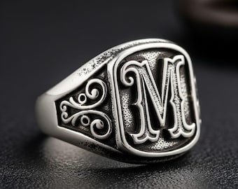 May include: A silver signet ring with the letter "M" in a bold, ornate font. The ring has a rectangular face with decorative scrollwork on the sides. The ring is made of a shiny, metallic material.