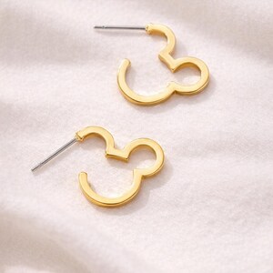 Könnte beinhalten: Ein Paar goldfarbene Creolen in Form eines Designs aus drei Kreisen. Die Ohrringe haben einen Steckerverschluss und werden auf einer weißen, strukturierten Oberfläche präsentiert. Das Design erinnert an eine berühmte Zeichentrickfigur.