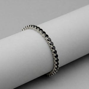 May include: A silver tennis bracelet featuring a row of round, black gemstones. The bracelet is presented on a white, curved surface, highlighting its design. The gemstones are evenly spaced and set in a silver metal, creating a continuous line.