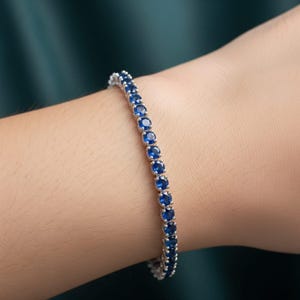 Pode incluir: Uma pulseira de prata com uma fileira de pedras preciosas redondas, azul escuro. A pulseira é usada em um pulso, mostrando as pedras brilhantes. O fundo é de cor verde-azulado escuro.