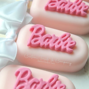 May include: Pink and white cake pops with the word "Barbie" in pink frosting on top. The cake pops are decorated with white bows and a sticker that says "Custom Treats & Company Gourmet Chocolate Treats".