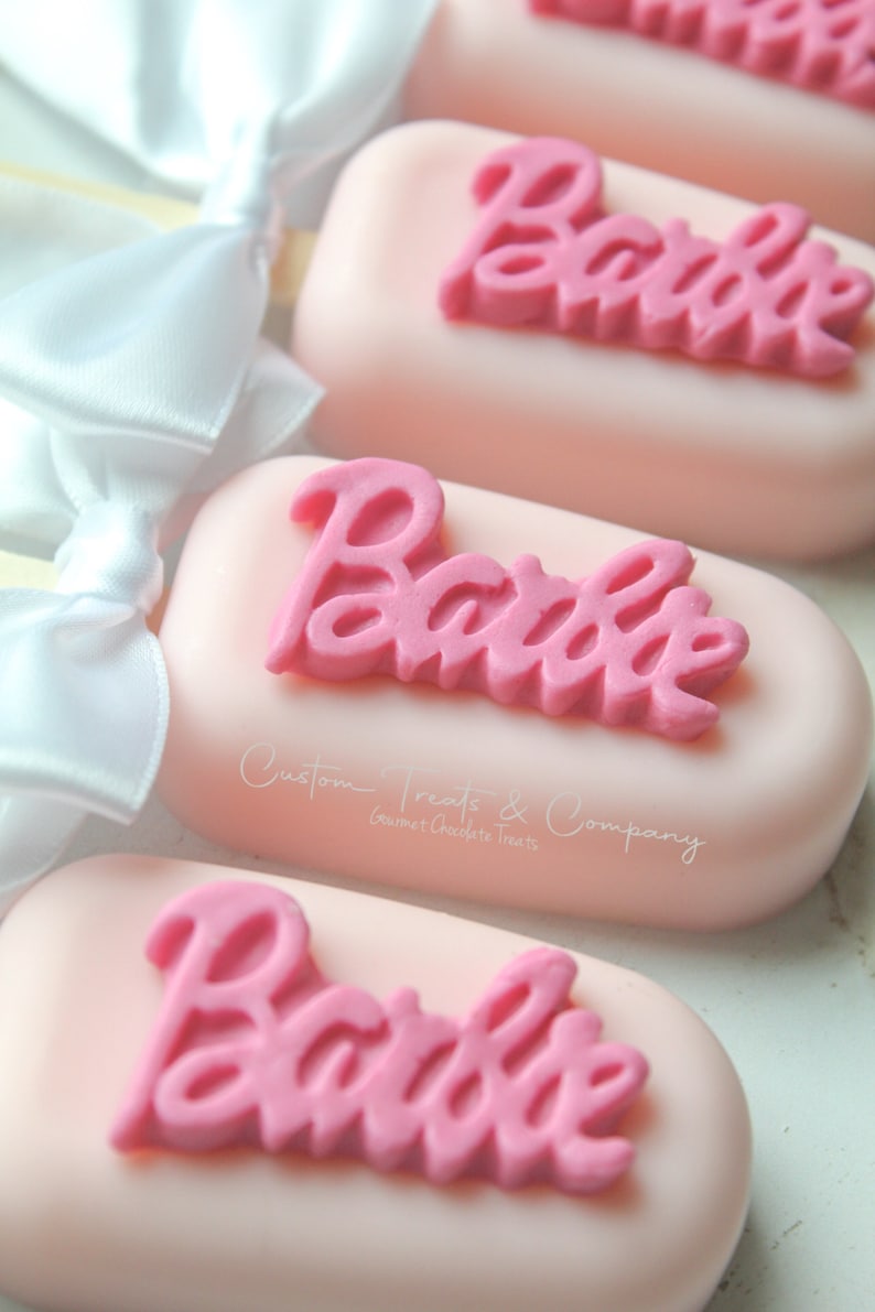 May include: Pink and white cake pops with the word "Barbie" in pink frosting on top. The cake pops are decorated with white bows and a sticker that says "Custom Treats & Company Gourmet Chocolate Treats".