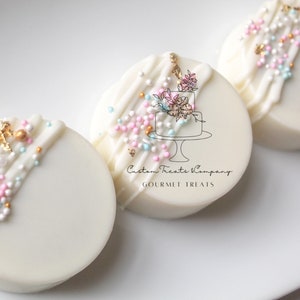 Puede incluir: Tres galletas cubiertas de chocolate blanco decoradas con glaseado blanco, chispas rosas y azules y detalles dorados. Las galletas tienen una pequeña cadena dorada y un logotipo que dice "Custom Treats Company Gourmet Treats".