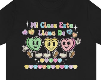 May include: Black t-shirt featuring a graphic of three candy hearts with faces. The hearts are green, yellow, and pink. The text "Mi Clase Esta Llena De Dulce Corazoncito" is printed below the hearts.
