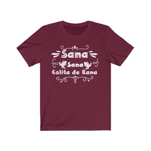 Op de afbeelding: Een maroon t-shirt met witte tekst die "Sana Sana Colita de Rana" luidt met twee kikkerillustraties.