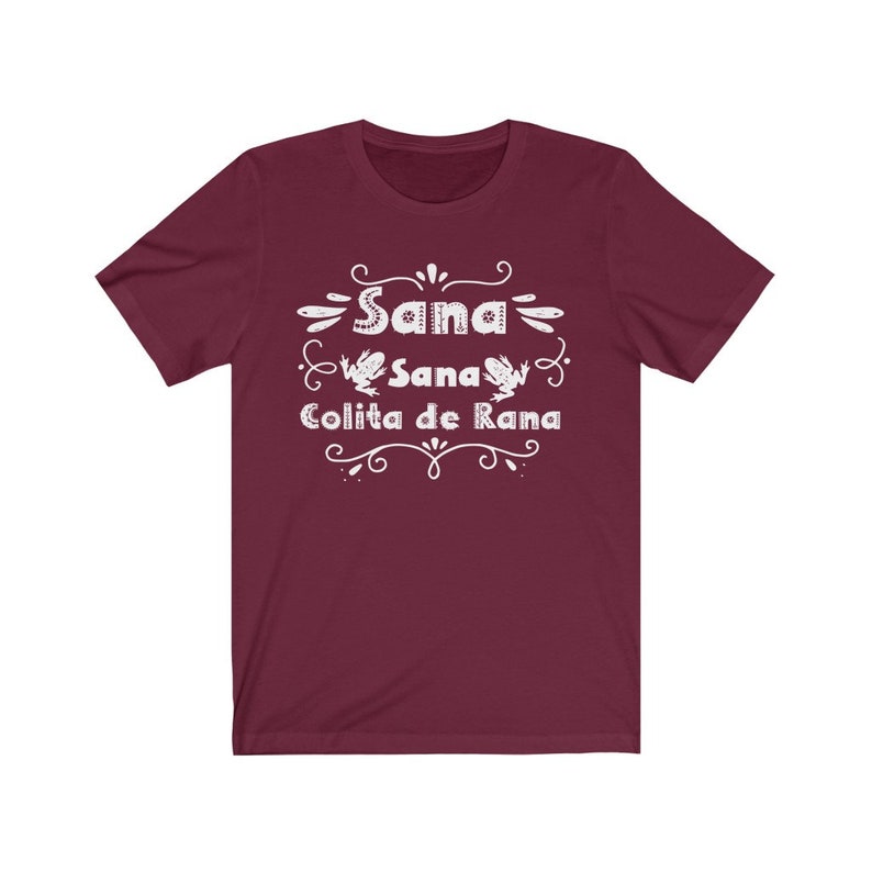 Op de afbeelding: Een maroon t-shirt met witte tekst die "Sana Sana Colita de Rana" luidt met twee kikkerillustraties.