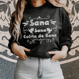 Op de afbeelding: Een zwart sweatshirt met witte tekst die "Sana Sana Colita de Rana" luidt met een decoratieve rand en twee kikkerillustraties.