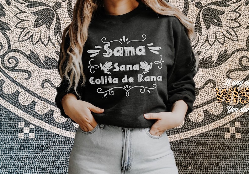 Op de afbeelding: Een zwart sweatshirt met witte tekst die "Sana Sana Colita de Rana" luidt met een decoratieve rand en twee kikkerillustraties.