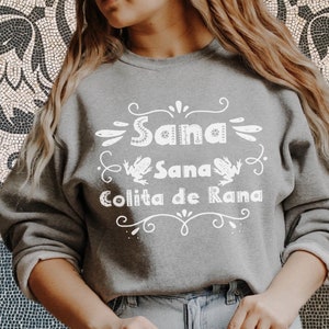 Op de afbeelding: Een grijs sweatshirt met een wit grafisch ontwerp met de tekst "Sana Sana Colita de Rana" met twee kikkers aan weerszijden van de tekst.