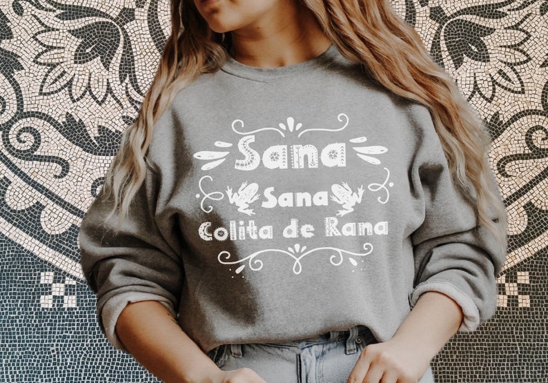 Op de afbeelding: Een grijs sweatshirt met een wit grafisch ontwerp met de tekst "Sana Sana Colita de Rana" met twee kikkers aan weerszijden van de tekst.