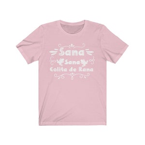 Op de afbeelding: Roze T-shirt met een wit grafisch ontwerp met de tekst "Sana Sana Colita de Rana" en twee kikkerillustraties.
