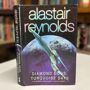 May include: A paperback book cover with a black background and a spaceship flying over a blue and purple planet. The title of the book is "Diamond Dogs, Turquoise Days" by Alastair Reynolds. The text "In the Revelation Space universe, anything can happen..." is printed below the title.