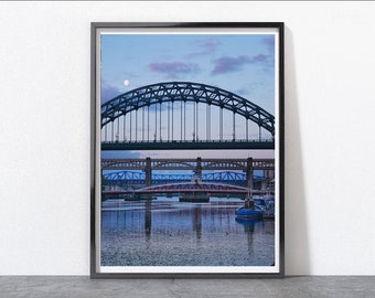 May include: A framed photograph of a bridge over a river at dusk. The bridge is made of steel and has a large arch. The river is calm and reflects the sky and the bridge. There is a small moon in the sky.
