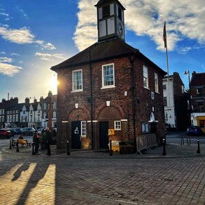 May include: A brick building with a clock tower and a flagpole flying a Union Jack flag. The building is in a town square with cobblestone streets and a few people walking by. The sky is blue with white clouds.
