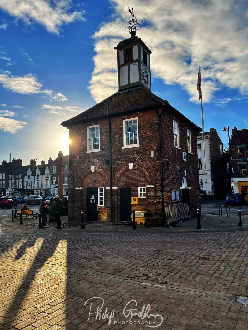 May include: A brick building with a clock tower and a flagpole flying a Union Jack flag. The building is in a town square with cobblestone streets and a few people walking by. The sky is blue with white clouds.