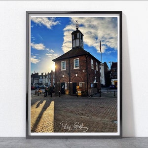 May include: A brick building with a clock tower and a blue sky with white clouds in the background. The sun is shining brightly and casting long shadows on the cobbled street.