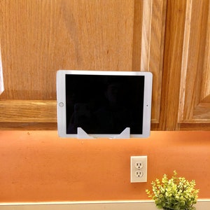 May include: A white tablet stand mounted on a wall above a white electrical outlet. The stand holds a black tablet in portrait mode. A small potted plant with green leaves sits on a light brown countertop.