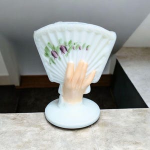 May include: A white glass fan-shaped figurine with a hand-shaped base. The fan is decorated with purple flowers and green leaves. The hand is painted with a flesh tone. The figurine has a round base.