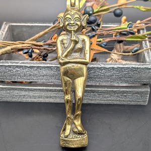 May include: A brass figurine of a smiling imp with pointed ears, a crown, and a finger to its lips. The imp stands on a base with engraved text. The figurine is detailed and has a vintage appearance.