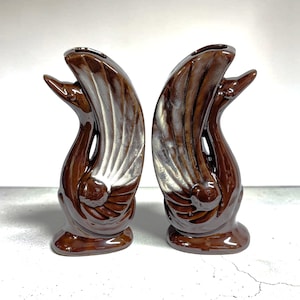 May include: A pair of glossy brown ceramic swan figurines, each featuring an elongated neck and stylized wing details with a metallic silver sheen on the feathers. The swans are positioned facing each other, with open tops that function as small vases.