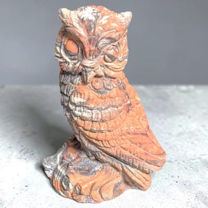Puede incluir: Una figura de búho tallada en piedra con patrones naturales de bandas en tonos terracota, óxido y gris. La escultura representa un búho con texturas de plumas detalladas, ojos cerrados y mechones en las orejas, posado sobre una base de piedra.