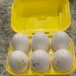 May include: A yellow plastic egg-shaped container holding six white egg-shaped salt and pepper shakers. Three shakers have cartoon faces. The container is open, revealing the shakers inside. The shakers have small holes on top.