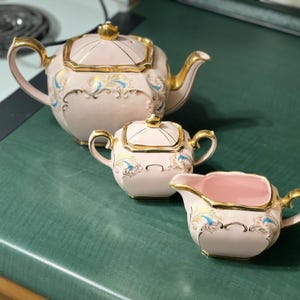 May include: A vintage tea set with a teapot, sugar bowl, and creamer. The set is pale pink with gold trim and decorative blue and gold accents. The teapot has a lid and a curved spout. The sugar bowl has a lid and handles. The creamer has a handle and a spout.