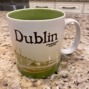 May include: White ceramic coffee mug with the word "Dublin" in brown lettering. The mug features a green interior and base, with a gold illustration of a building. The Starbucks Coffee logo is also visible.