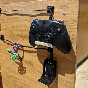 Könnte beinhalten: Ein schwarzer Xbox-Controller ist an einer Wand mit einer schwarzen Ladestation befestigt. Der Controller ist über ein schwarzes Kabel mit der Ladestation verbunden. Die Ladestation ist mit einem schwarzen Haken an der Wand befestigt.
